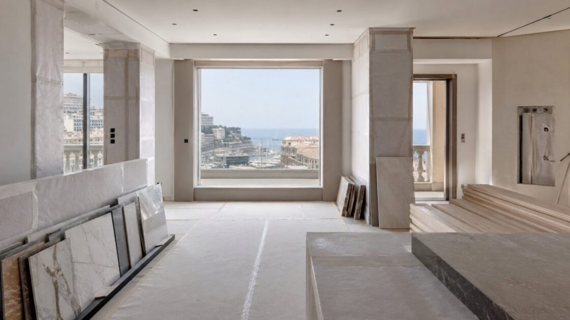 Chantier d’un appartement : dalles de marbre en attente de pose, sols protégés, baie ouverte sur un port méditerranéen