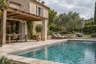 Piscine bordée de pierre claire devant une façade en pierre et une terrasse sous pergola en bois